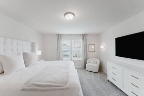 A modern, white-themed bedroom with a large bed, a bedside table with a lamp, a chair, a TV, and a window with curtains. - Luxury 5bedroom Home, Pool, Near Disney (Clermont)