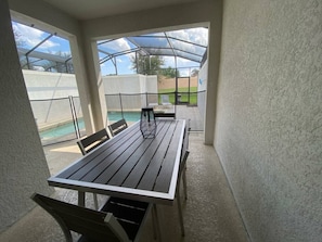 A covered patio with a table and chairs, a pool in the background, and a clear view of the sky. - Your Orlando Home With Private Pool (Davenport)