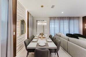 Een moderne eetruimte met een lange tafel gedekt voor zes personen, een kroonluchter en een bank op de achtergrond.