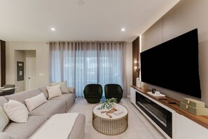 Ein modernes Wohnzimmer mit großem Flachbildfernseher, einem beigen Sofa, zwei dunklen Sesseln und einem runden Couchtisch.