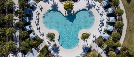 An aerial view of a heart-shaped swimming pool surrounded by lounge chairs and umbrellas, with palm trees and a pathway around it.