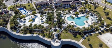 An aerial view of a resort with a central swimming pool, a bridge, and surrounding greenery.