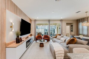 A modern living room with a flat-screen TV, a coffee table, sofas, and a dining area with chairs. - Luxury 6BR Home With Pool Solara Resort (Kissimmee)