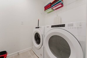 A laundry room with two white washing machines, a shelf with folded towels, and a hexagonal wall decoration. - Luxury 6BR Home With Pool Solara Resort (Kissimmee)