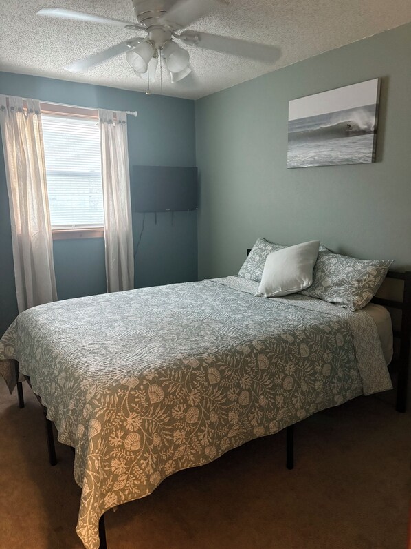Un dormitorio con una cama, ventilador de techo, ventana con cortinas y televisor montado en la pared.