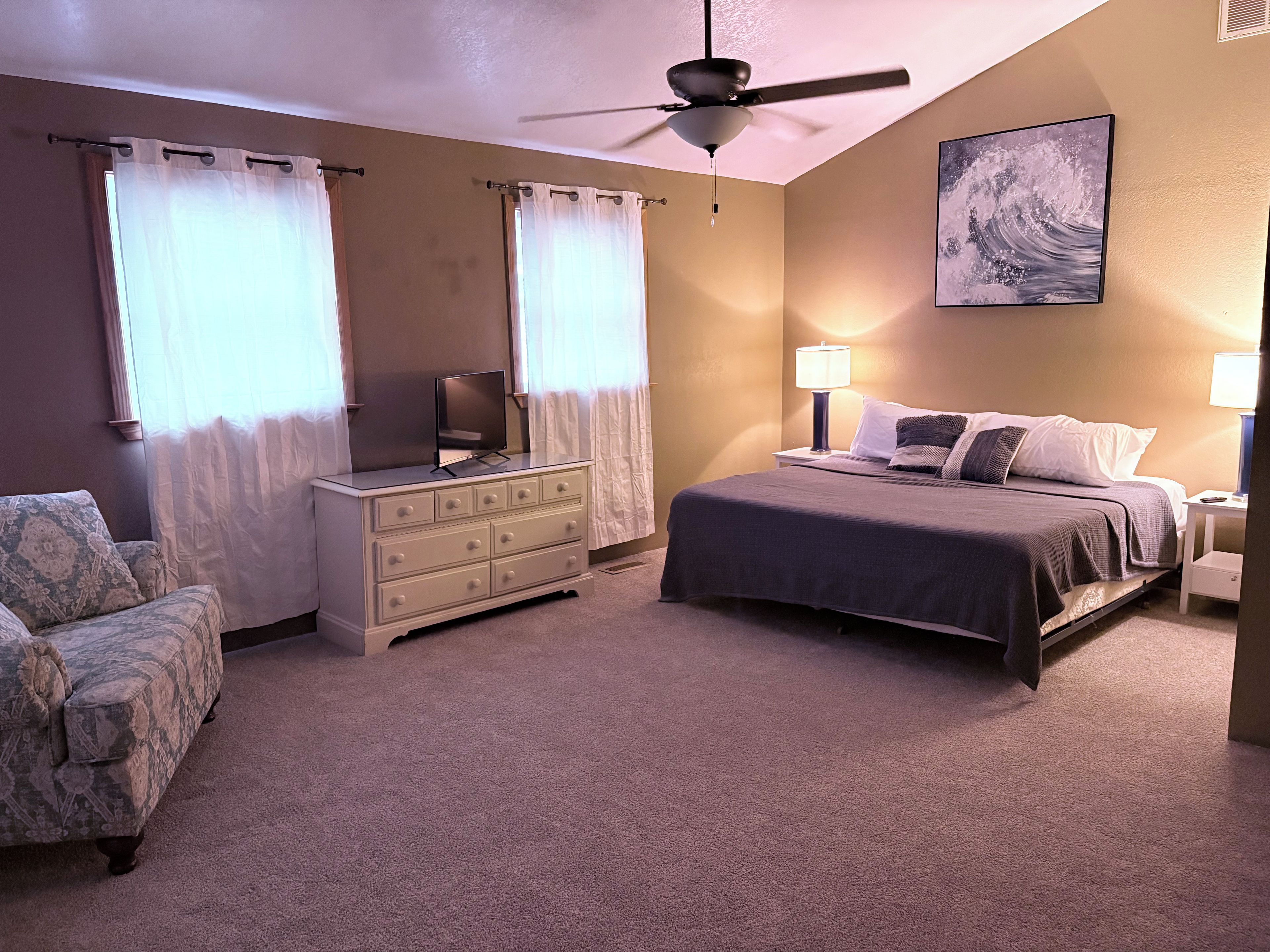 A bedroom with a bed, dresser, chair, ceiling fan, and artwork on the wall.