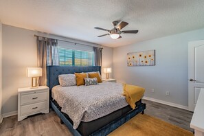 Una camera da letto con un letto, comodini, una ventola a soffitto, una finestra con tende e un quadro appeso al muro.