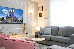 A modern living room with a grey sofa, pink armchairs, a flat-screen TV, and a small round table. - Disney 4BR Home w/ Pool + Themed Bedrooms (Kissimmee)
