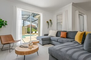 A modern living room with a grey sectional sofa, a wooden coffee table, and a brown chair. There is a glass door leading to a pool area. - Storey Lake 4BR w/ Pool + Disney-themed Rooms (Kissimmee)