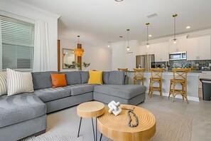 A modern living room with a grey sectional sofa, wooden side tables, and a kitchen area with bar stools and a refrigerator. - Storey Lake 4BR w/ Pool + Disney-themed Rooms (Kissimmee)