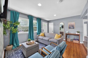 A modern living room with a grey sofa, teal curtains, a small potted plant, and a wooden floor. - Lovely Truist Retreat | Cozy 4 Bed with Fire Pit (Marietta)