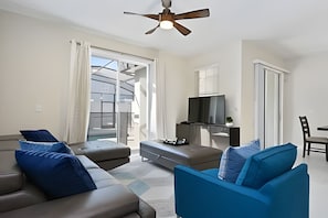 A modern living room with a sofa, ottoman, and a TV on a stand. There is a ceiling fan, a sliding glass door, and a dining area with a chair. - Charming 4BR w/ Pool Near Disney - Storey Lake (Kissimmee)
