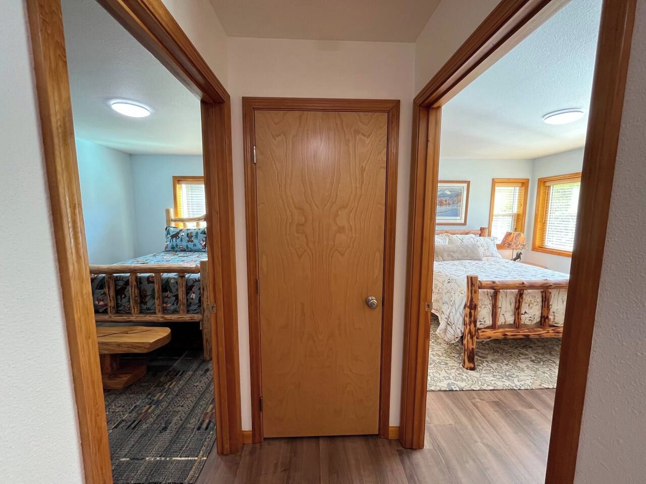 Un couloir mène à une chambre dotée d’une porte en bois, d’un lit recouvert d’une couvre-lit à motifs et d’une fenêtre avec des stores.