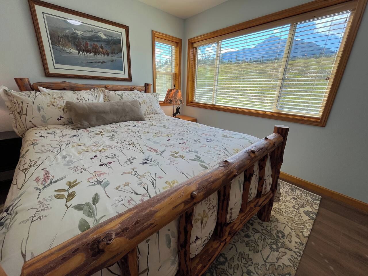 A bedroom with a large bed, wooden headboard, floral bedding, a painting on the wall, and a view through the window.
