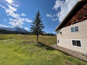 Une maison avec un toit en pente et une antenne parabolique, entourée d’un champ verdoyant et avec une montagne en arrière-plan.