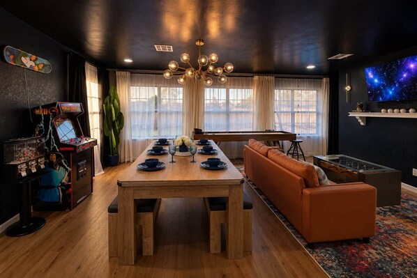 A room with a wooden dining table set for four, a sofa, a foosball table, arcade games, and a large TV screen.