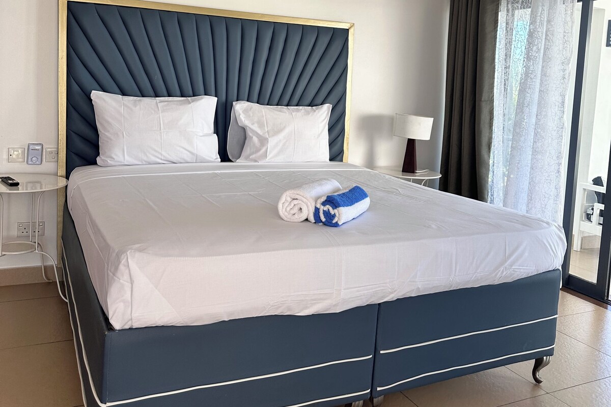 Quarto de hotel com cama, mesa de cabeceira, abajur e vista para o exterior pela janela.