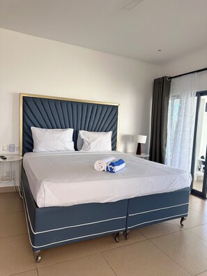 Quarto de hotel com cama, mesa de cabeceira, abajur e vista para o exterior pela janela.