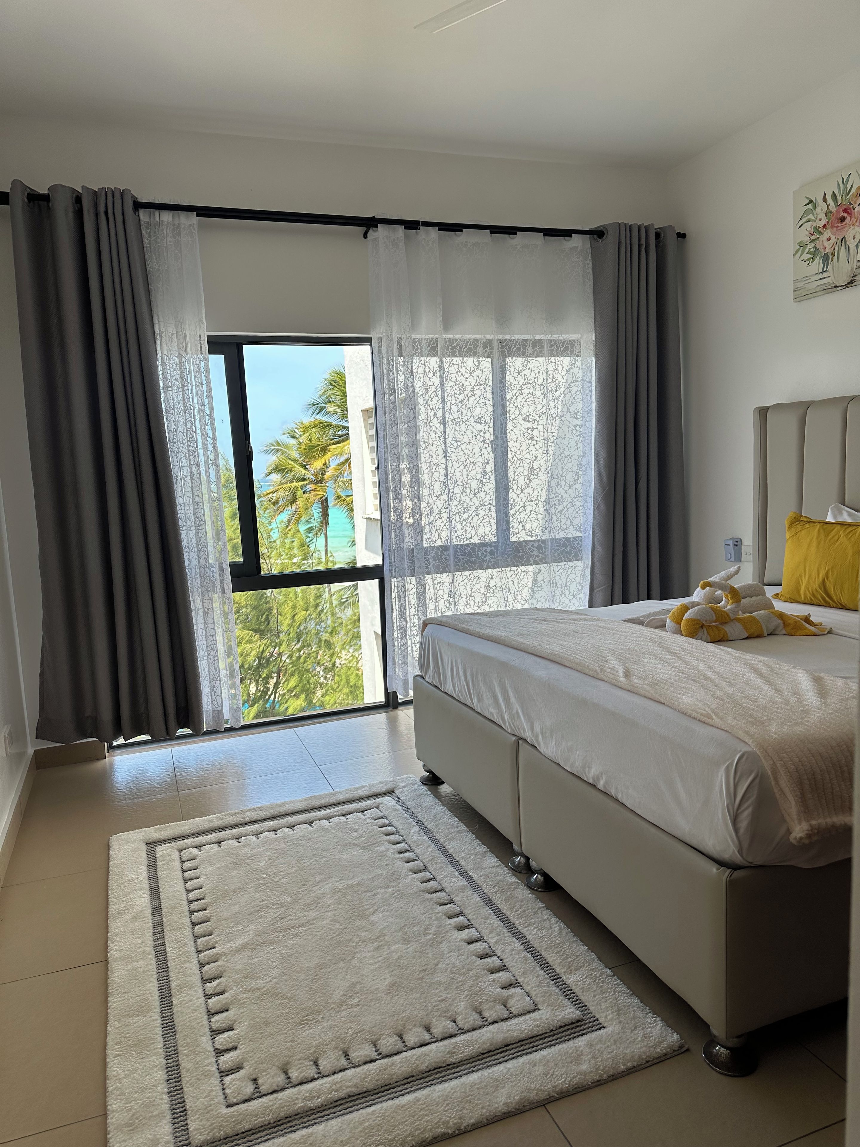 Un dormitorio con cama, ventana con cortinas, vista a palmeras y una alfombra blanca.