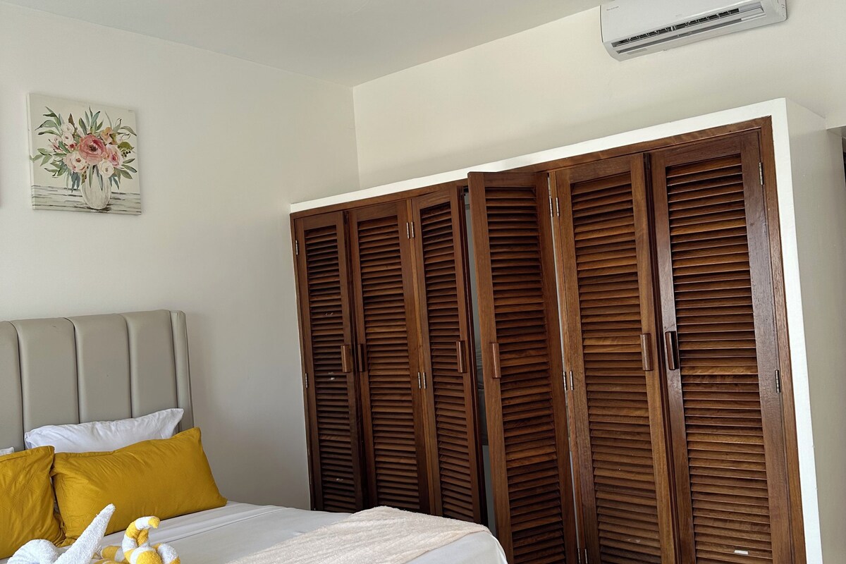 Quarto com cama, ventilador de teto, ar condicionado, guarda-roupas de madeira e um quadro na parede.