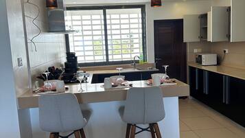 A modern kitchen with a central island, two bar stools, pendant lighting, and a view through a large window.