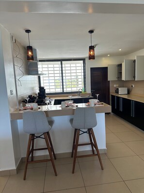 Cozinha moderna com ilha central, dois banquetes, iluminação pendente e vista através de uma grande janela.