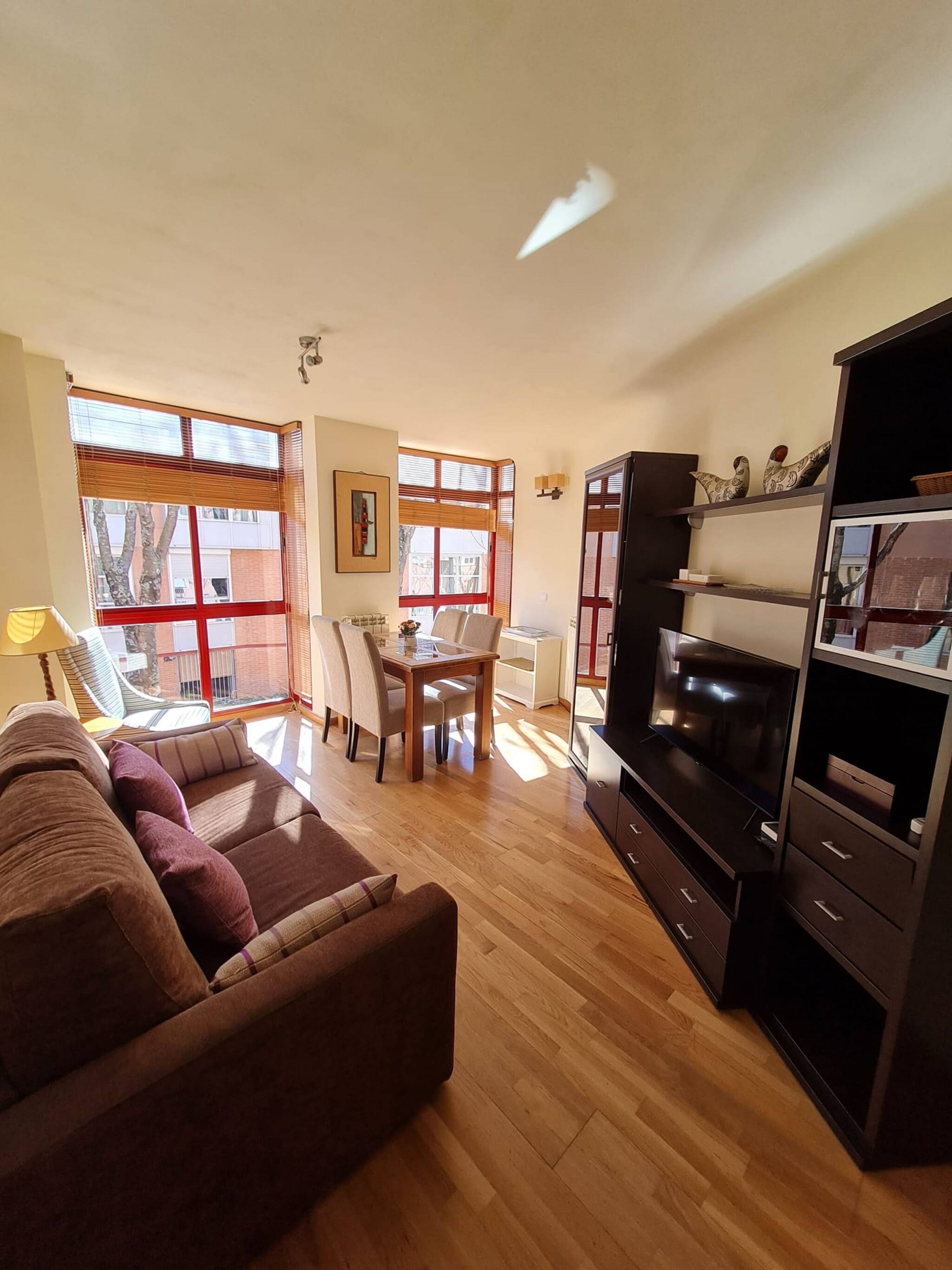 Una sala de estar con sofá, televisor, mesa de comedor con sillas y un amplio ventanal con persianas.