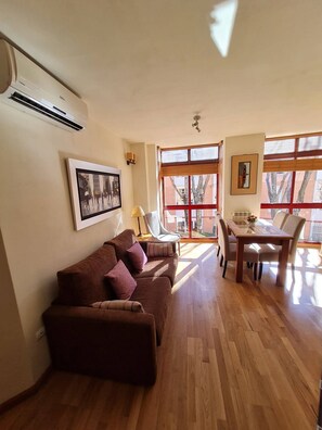A living room with a brown sofa, a glass-top dining table with chairs, and a large window with blinds. - Great Apartment in Madrid (Madrid)