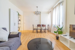 A modern living room with a sofa, a dining table with chairs, a chandelier, and a TV on a stand.