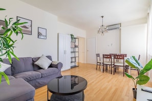 A modern living room with a grey sofa, a black coffee table, wooden flooring, a dining area with chairs, a white door, and a plant. - Spacious Room in Madrid (Madrid)