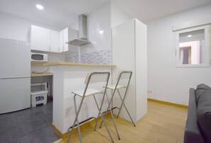 A compact kitchen with white cabinets, a microwave, and a wooden countertop. There are two foldable chairs and a window with blinds. - Apartment in Fuente del Berro (Madrid)