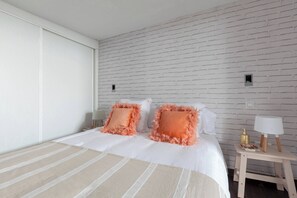 A hotel room with a white brick wall, a bed with striped bedding, two pillows with fringe, a bedside table with a lamp, and a small nightstand with a bottle. - Cool urban loft at Madrid's heart. Malasaña. (Madrid)
