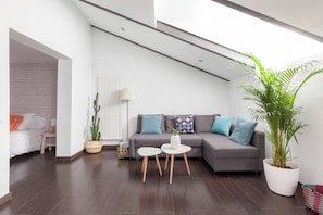 Ein modernes Wohnzimmer mit einem grauen Ecksofa, zwei weißen Beistelltischen, einem Blumentopf und einer Wand mit Ziegeloptik.