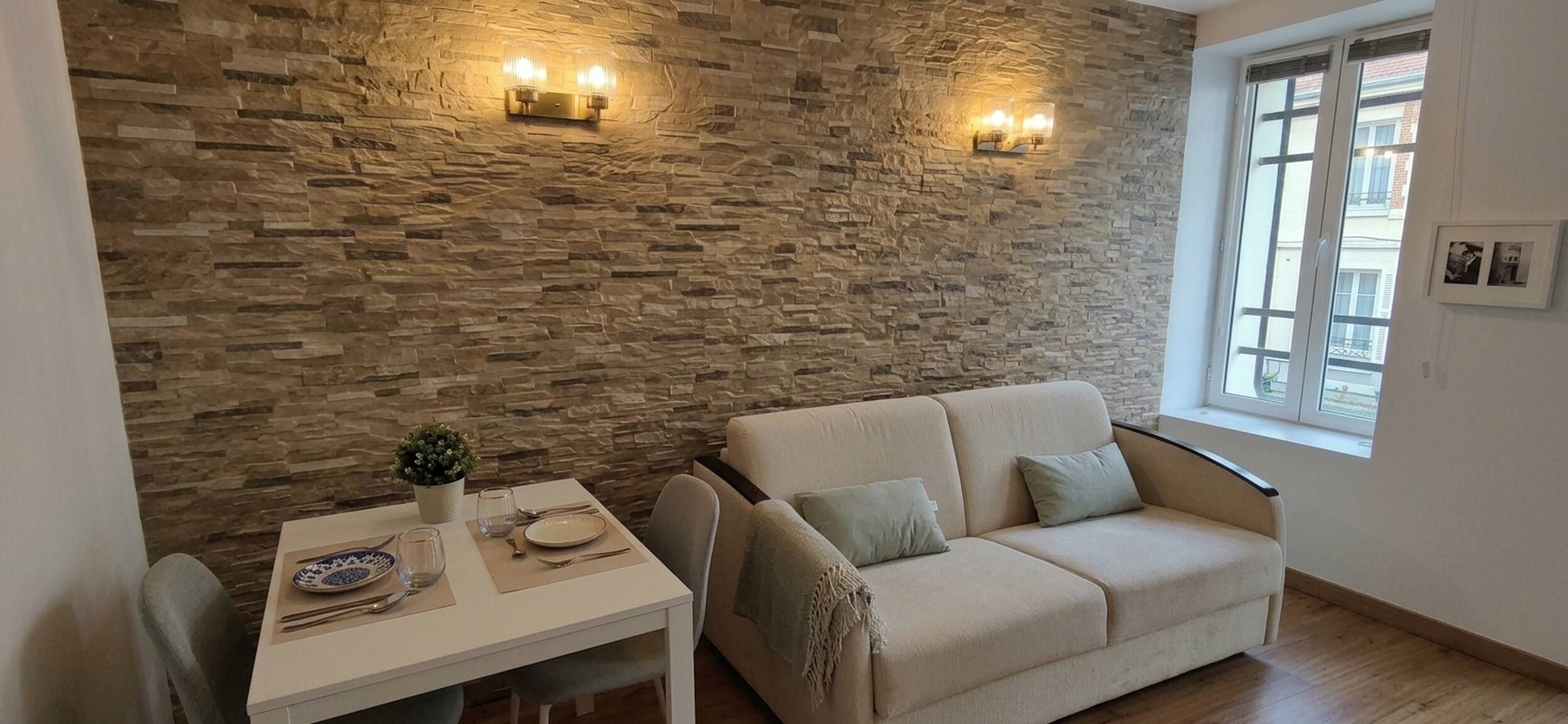 Una sala de estar con una pared de piedra, un sofá, una mesa de comedor con una planta y una ventana.