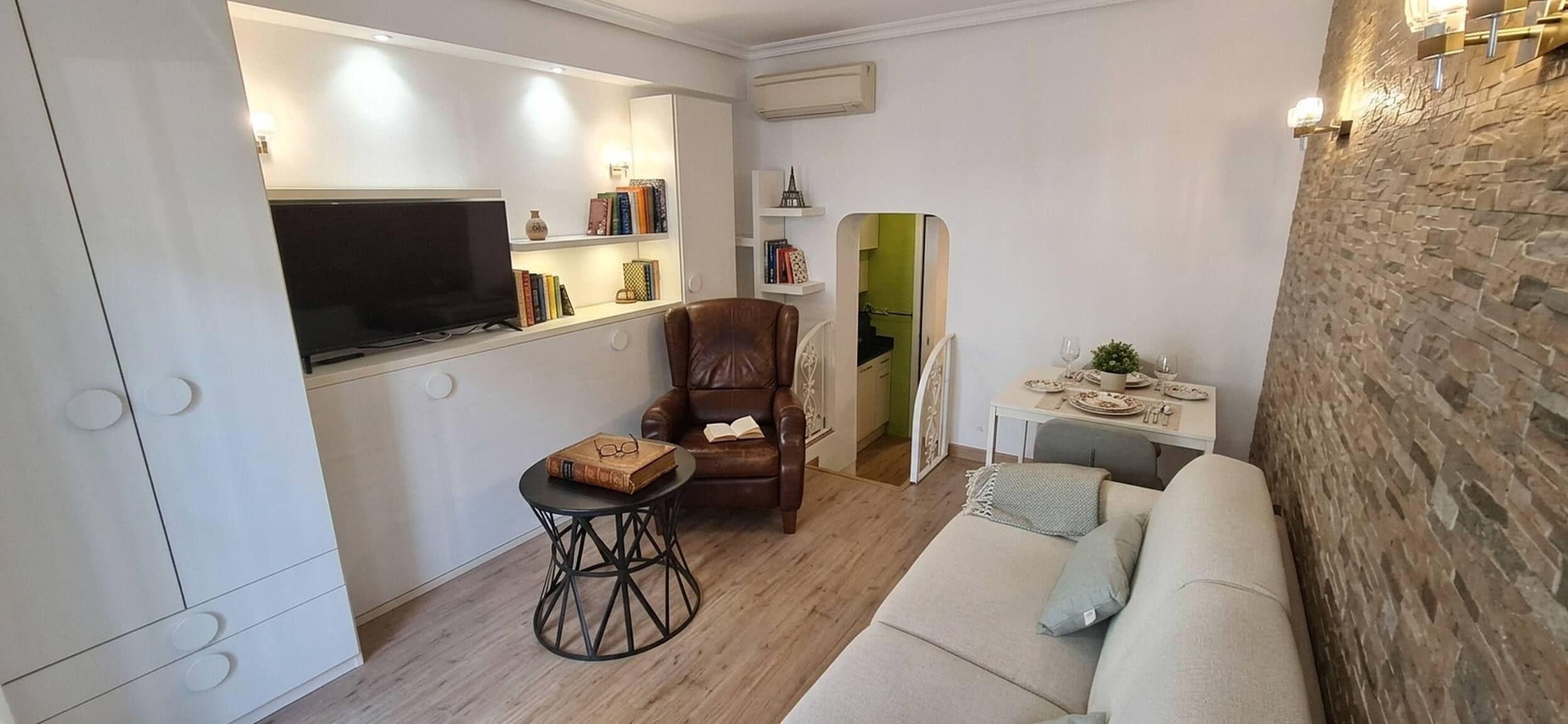 Un espacio habitable compacto con un sofá, una pequeña mesa, un sillón de cuero, un televisor y una estantería.