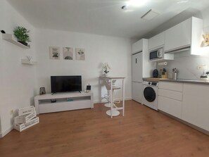 Cozinha moderna com micro-ondas, pia e máquina de lavar. Há uma TV em um suporte branco e uma pequena mesa com um vaso de planta.