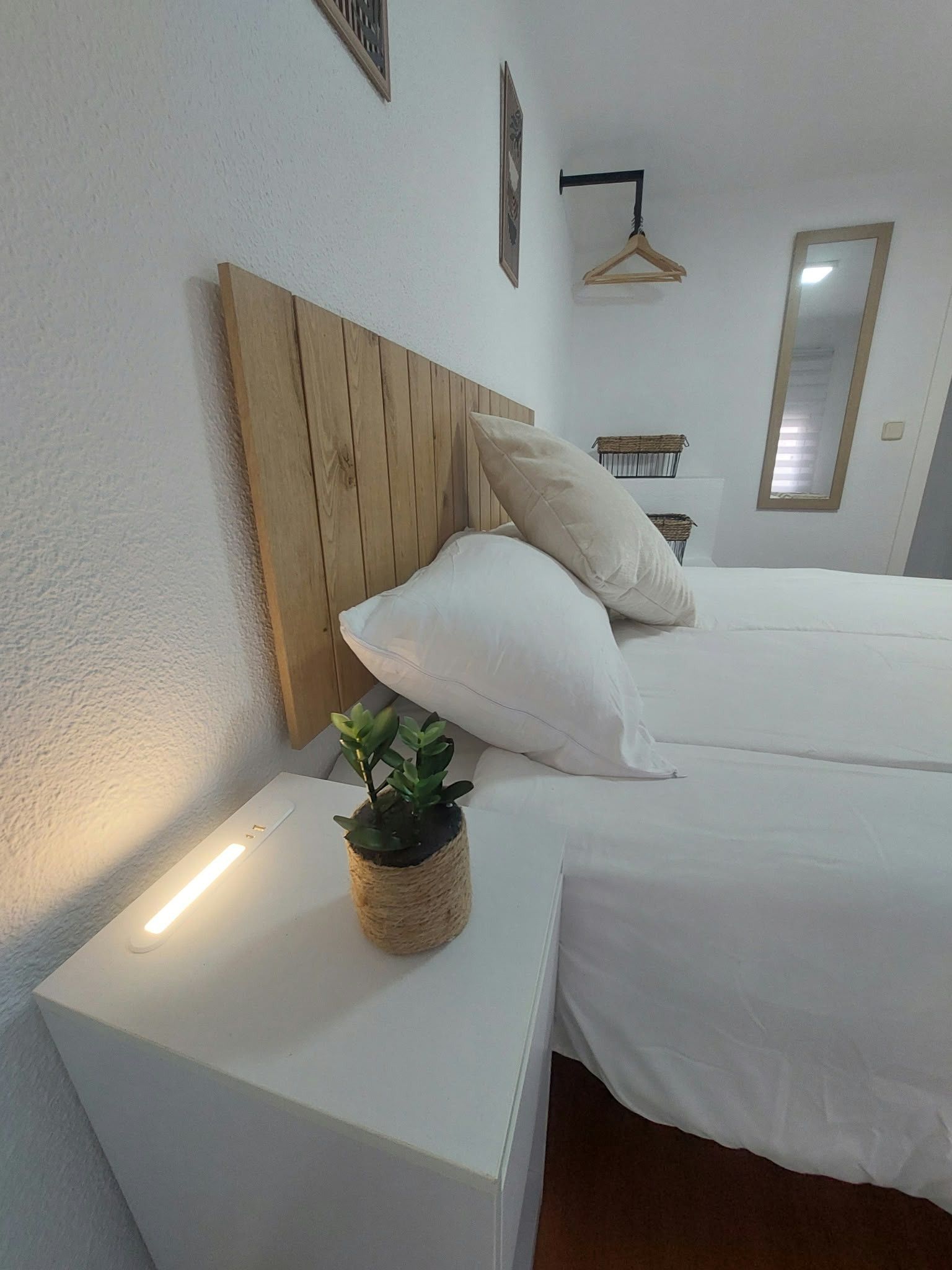 A bedroom with a wooden headboard, two white beds, a bedside table with a potted plant, and a hanging light fixture.