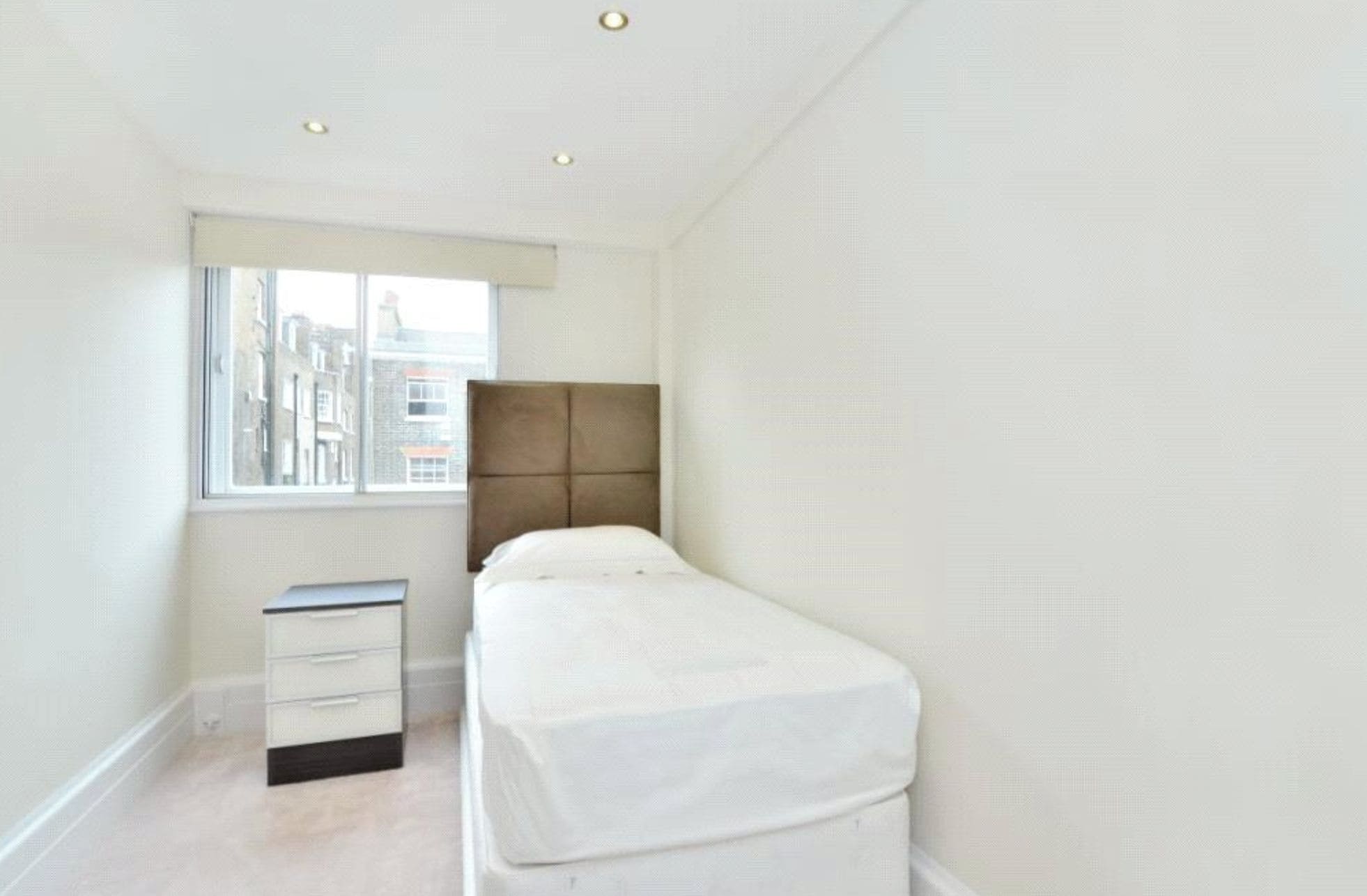 A small, single-bed room with a window, a bedside table, and a wardrobe.