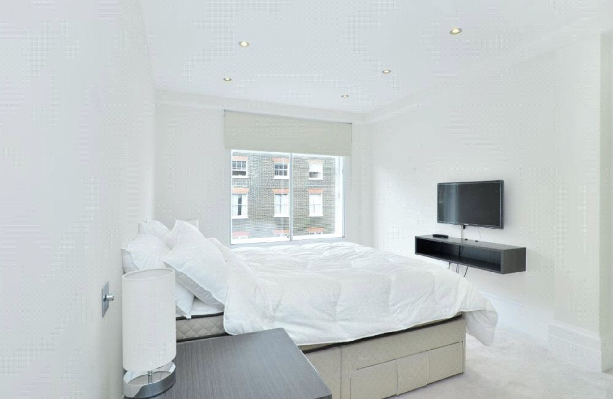A modern bedroom with a bed, a TV mounted on the wall, and a window with a view of buildings.