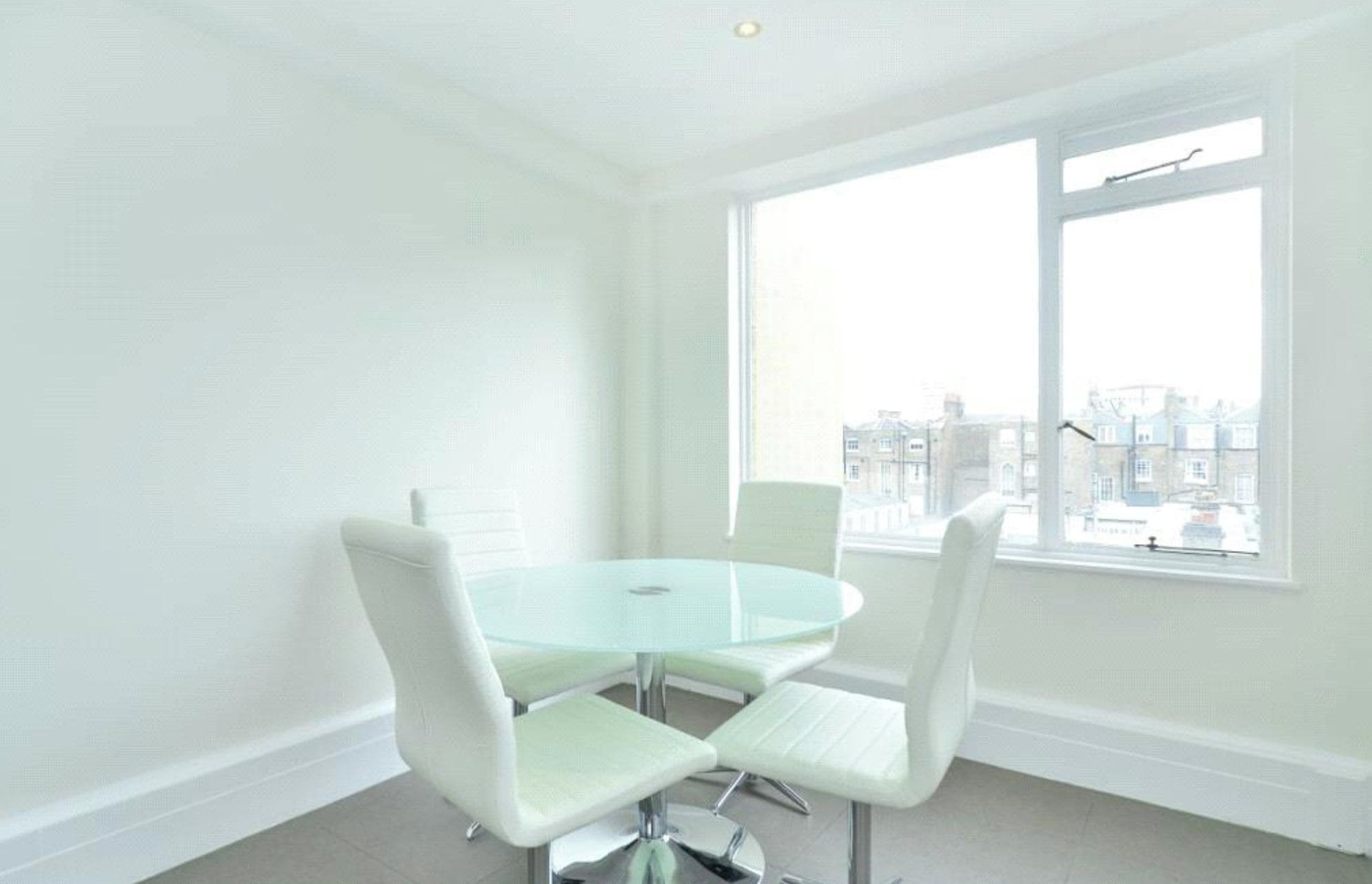 A room with a round glass table and four white chairs, a large window with a view of buildings, and white walls.