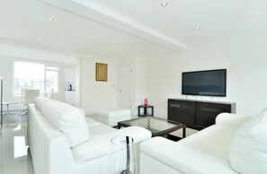 Un salon moderne avec des meubles blancs, une table basse en verre et un téléviseur à écran plat fixé au mur.