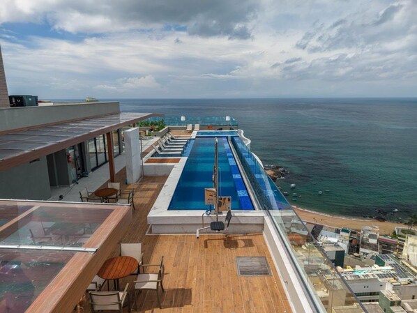 A rooftop pool with a view of the ocean, wooden deck, and lounge chairs.