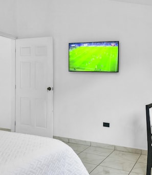 Una habitació amb un llit, una porta blanca, una televisió penjada a la paret que mostra un esdeveniment esportiu i un sòl de rajoles.