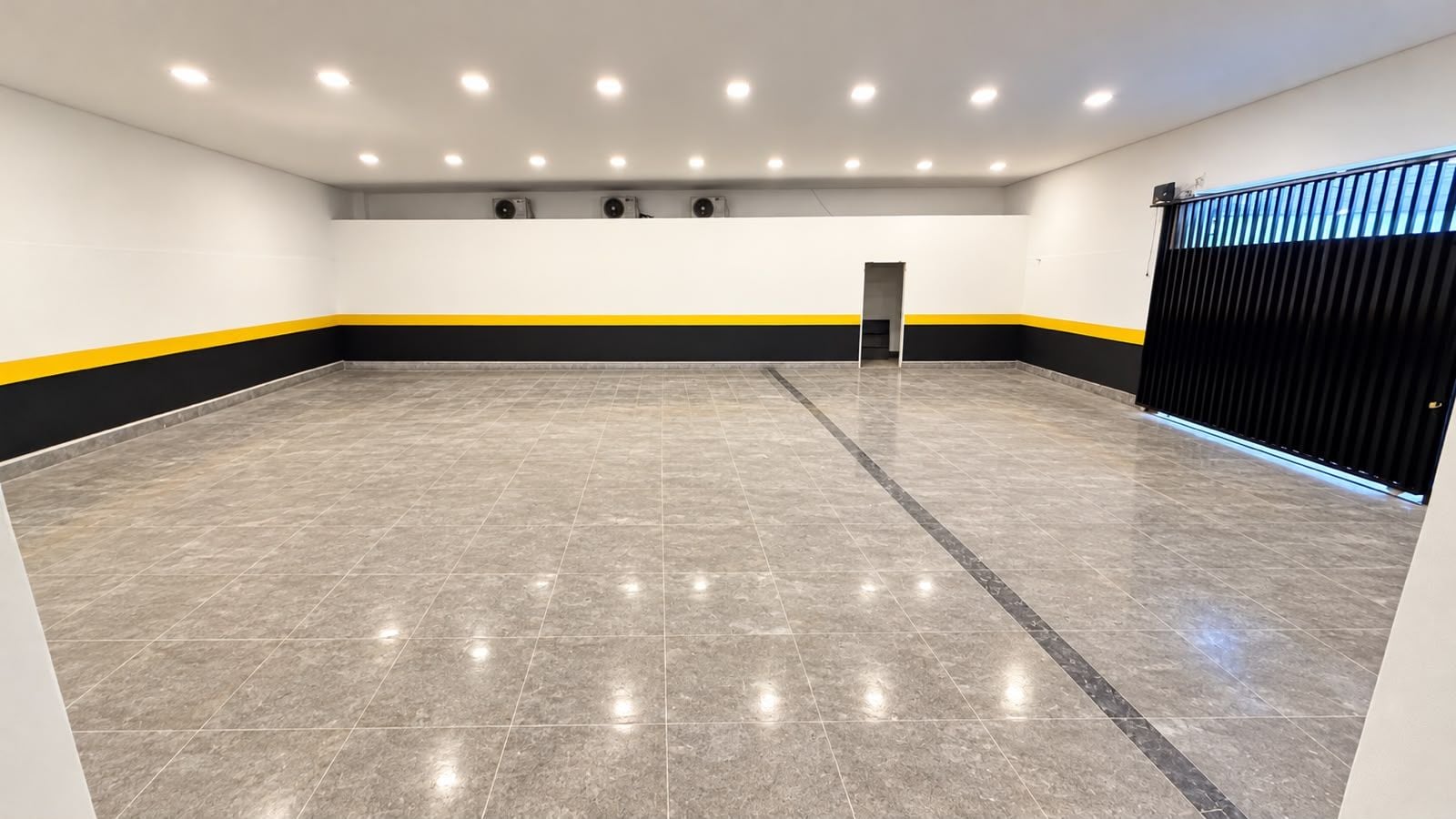 A spacious indoor area with a tiled floor, a black and yellow stripe along the walls, and a black gate on the right.