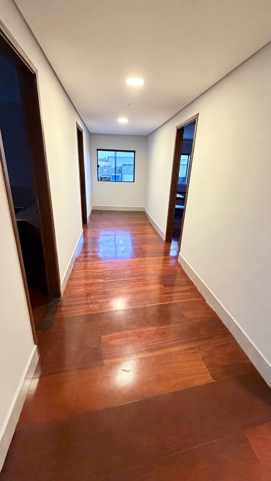 Un pasillo con piso de madera, paredes blancas y un ventanal al fondo.