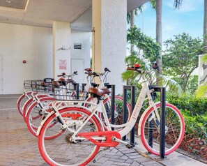 A row of bicycles with red tires parked in an outdoor area with palm trees and a building in the background. - BW Resort 2BR APT - Beach Access - Fitness Center (Hallandale Beach)