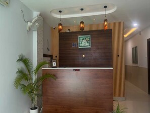 A reception desk with a wooden finish, a framed artwork above it, and a potted plant to the left. - Deluxe Hotel C&D Stay (Indore)