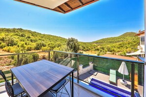 A balcony with a table and chairs, a pool area with an umbrella, and a view of a green landscape. - Villa Çınar-kaş Antalya (Kaş)