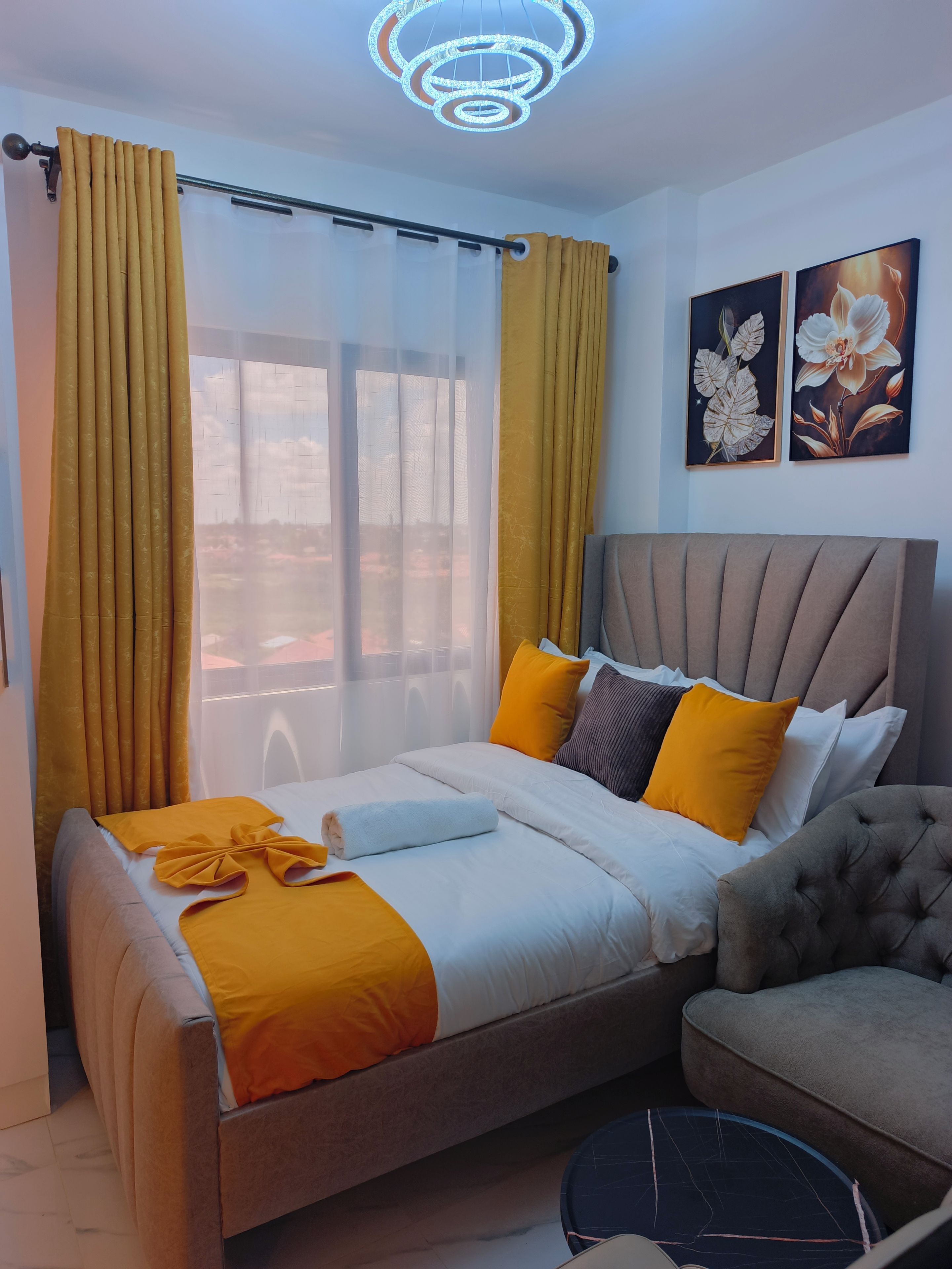 A bedroom with a bed, a sofa, a chair, a ceiling light, and two wall-mounted paintings.