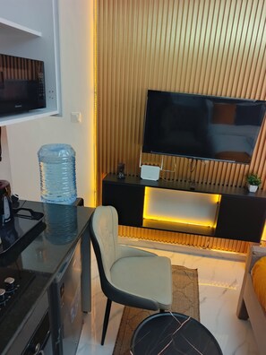 A modern living room with a flat-screen TV, a water dispenser, a sofa, and a dining area with a coffee machine. - Cozy Studio apartment near JKI Airport and SGR in Nairobi perfect for your stay (Nairobi)